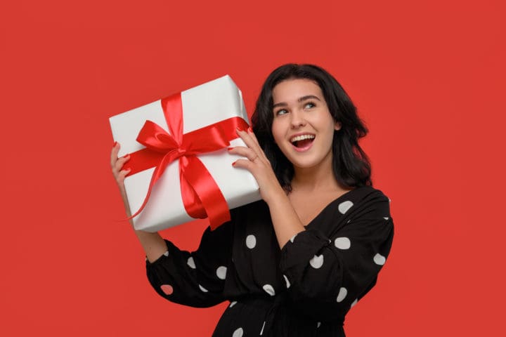 Brunette woman in a black dress smiling holding Christmas giftbox of white color with red ribbon