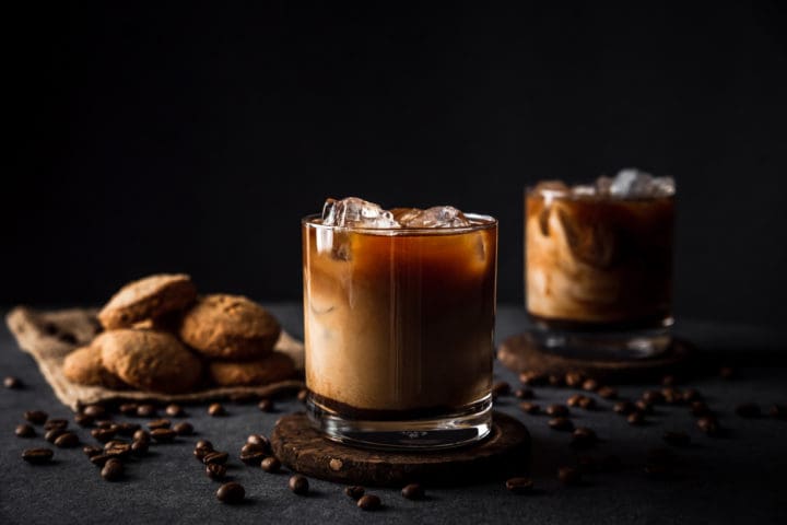 A horizontal photo of rocks glasses with cold iced coffee with milk, coffee beans around, cookies, dark background, deep shadows