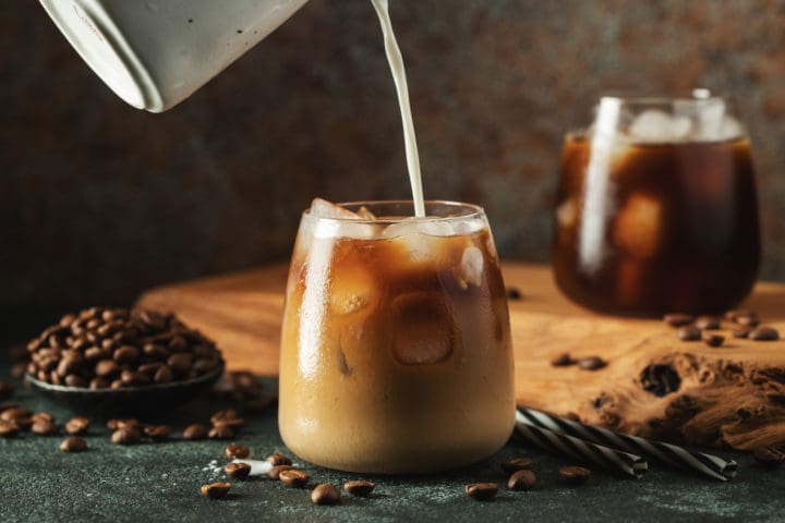 Ice coffee in a tall glass with cream poured over, ice cubes and beans on a dark concrete table