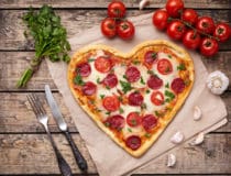 Heart shaped pizza for Valentines day with pepperoni, mozzarella, tomatoes, parsley and garlic on vintage wooden table background Food symbol of romantic love