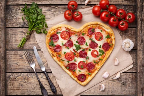 Heart shaped pizza for Valentines day with pepperoni, mozzarella, tomatoes, parsley and garlic on vintage wooden table background Food symbol of romantic love