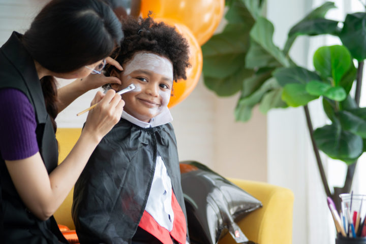 Mother and son in Halloween costumes having fun with makeup, mother paints a pattern on her son's face with a paintbrush, boy smiling and looking at camera, happy halloween together